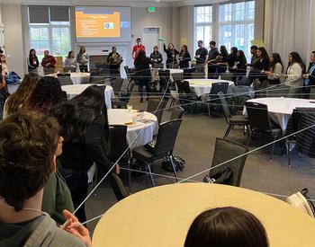 Latinx teenagers are in a large circle in a conference room in an icebreaker activity that involves holding lines of string that criss-cross the room.