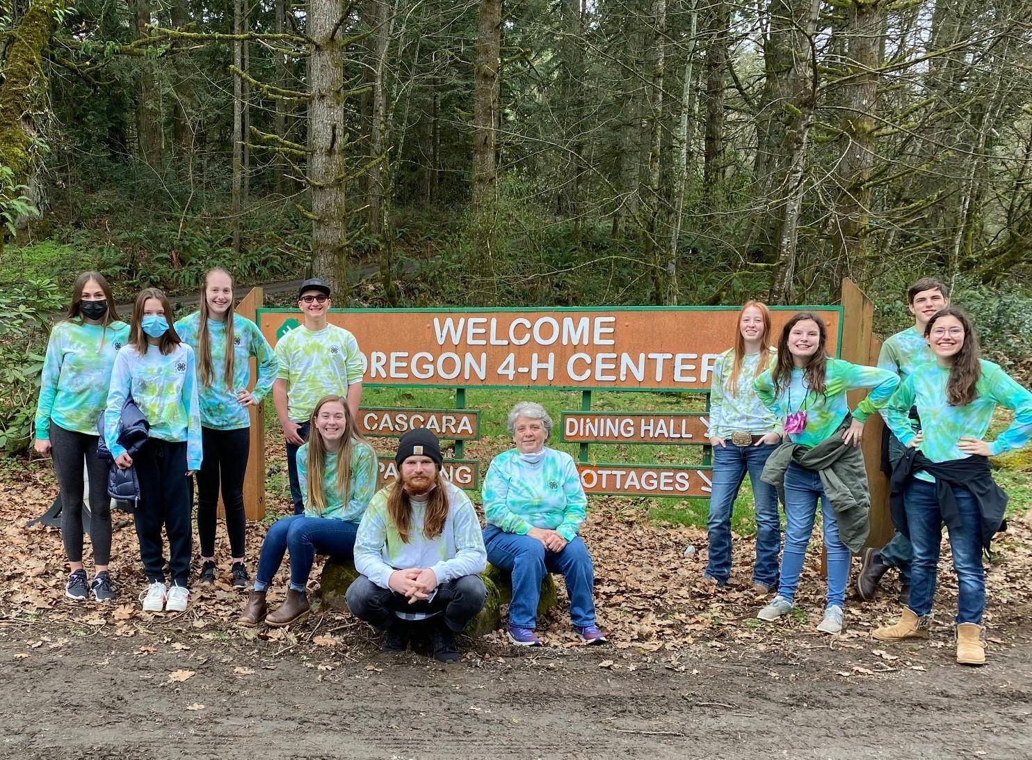 A group of 4-H youth participants and leaders stand outdoors in front of a sign reading "Welcome Oregon 4-H Center"