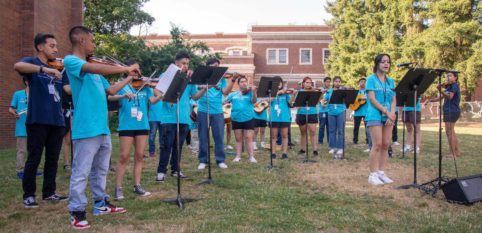 Teenagers wearing blue shirts are playing violins, trumpets and acoustic guitars at an outdoor concert.