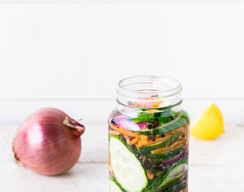 A jar of cucumbers and carrots is pictured with a whole red onion, a half lemon and a sprig of a fresh herb.