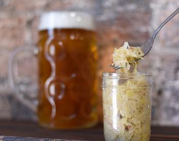 A jar of sauerkraut is shown with someone displaying a forkful of the sauerkraut over the jar. In the background is a large mug of beer.