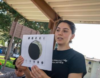 A woman wearing a black T-shirt is holding up a piece of paper with the center cut out to show the phases of the moon.