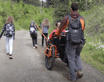 Five youths walk on a nature trail while one pushes a a wheelchair used by a girl.