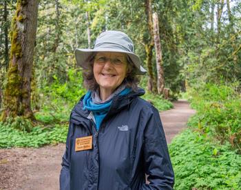 A woman is posing for a photo on a trail in a forest. She's wearing hiking apparel. and a floppy hat.