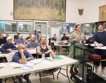 Incarcerated students sitting at tables in a classroom in the Pregon State Penitentiary are smiling as they look at a screen.