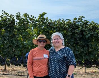 Two woman stand next to each other smiling. Behind them there are blue skies and vineyards.