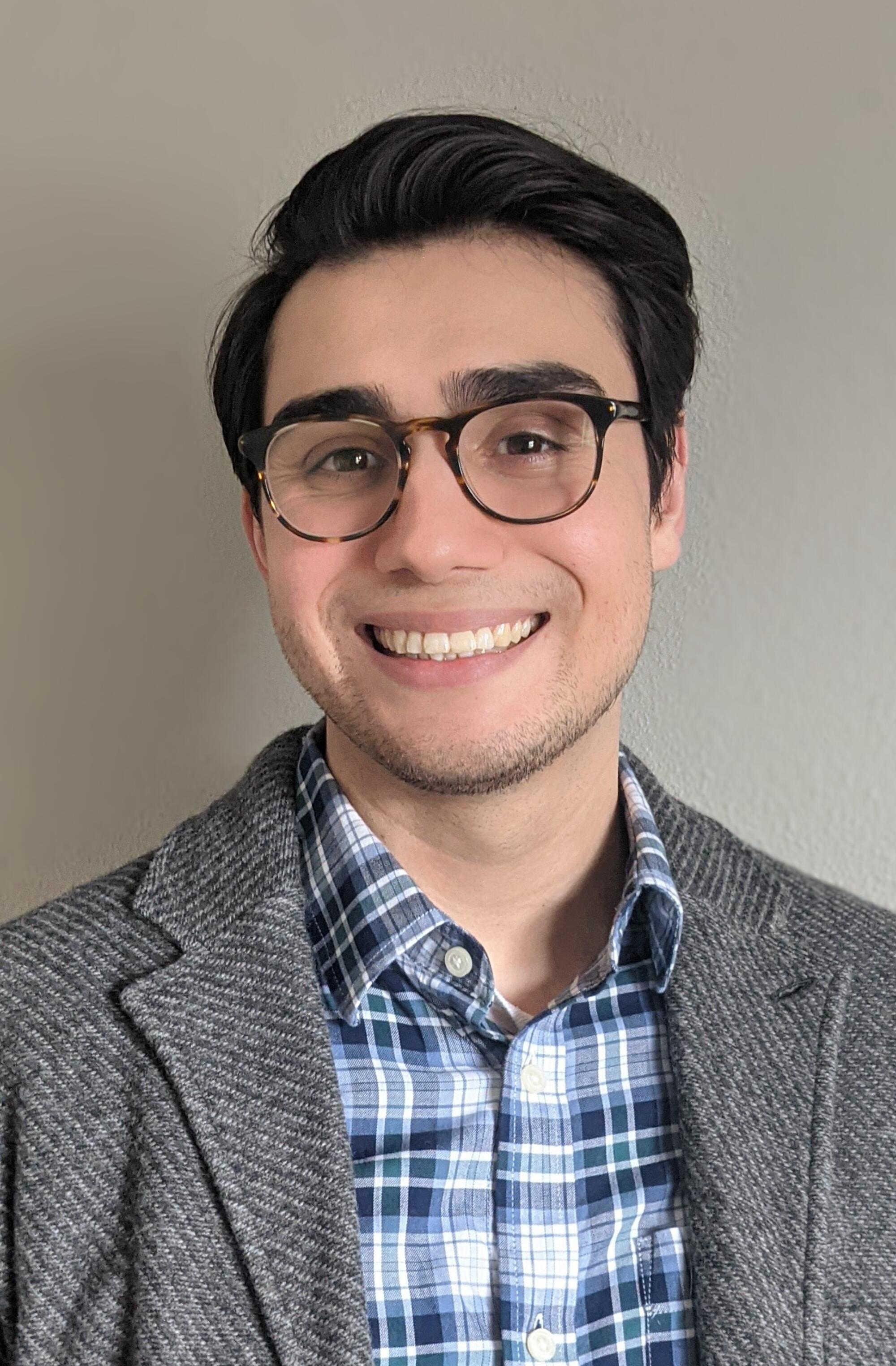 A smiling person wearing glasses, a plaid shirt and a blazer stands against a neutral background in a professional portrait.