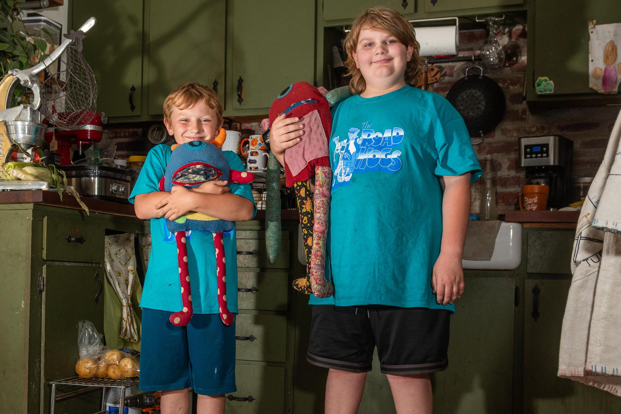 Ben LaMont and his brother Wyatt stand in their kitchen, smiling toward the camera while holding handmade “worry monster” crafts, with tools and materials visible on shelves behind them.