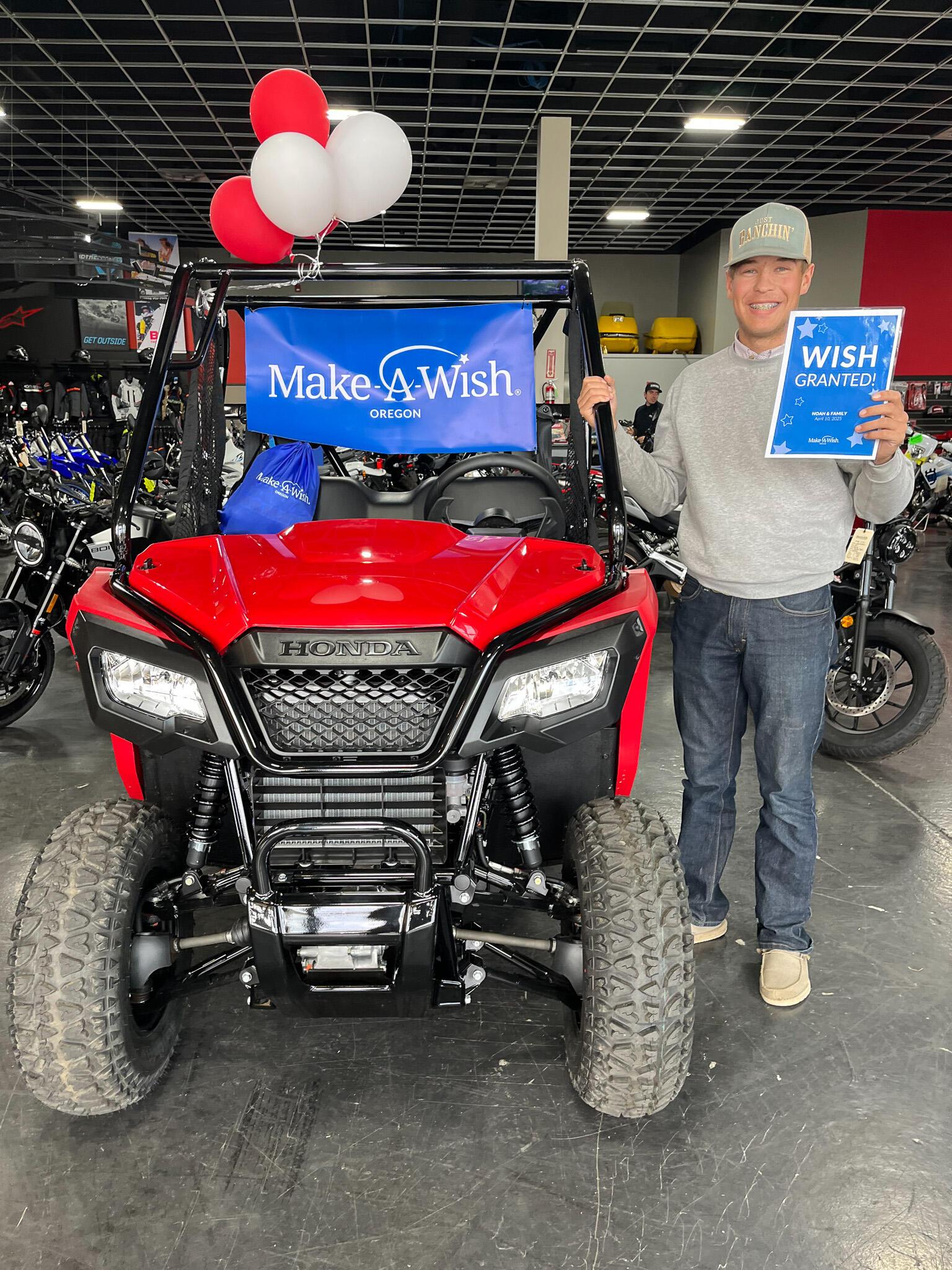 Noah Baker stands indoors beside his new red Honda side-by-side vehicle, holding a blue “Wish granted” sign in front of a Make-A-Wish Oregon banner, with motorcycles and equipment visible in the background.