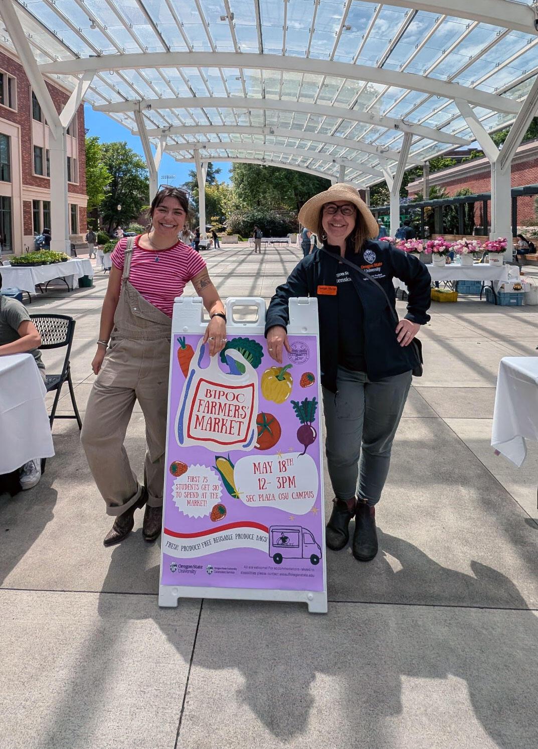 Ainsley McCollum and Teagan Moran stand beside a sign for the BIPOC Farmers Market in SEC Plaza at Oregon State University, under a glass canopy surrounded by produce and flower vendors.