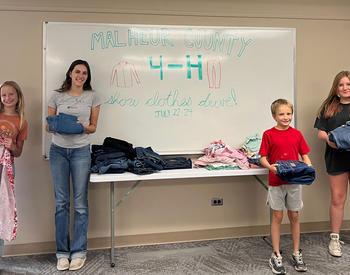 Four youths stand beside a table stacked with folded clothing during a Malheur County 4-H clothes drive, with a handwritten sign on the whiteboard behind them celebrating the donation effort.
