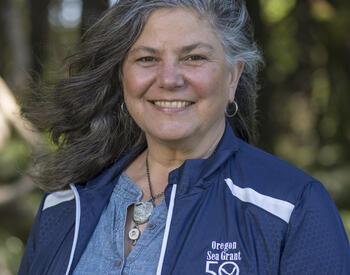 A woman with long gray hair smiles outdoors while wearing a blue jacket with the Oregon Sea Grant 50 Years logo.