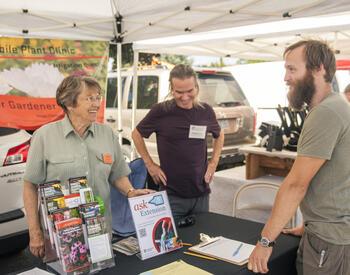 three people talking at an outdoor information booth under a canopy; two volunteers stand behind a table with brochures and posters while a visitor listens and smiles.