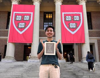 Yahir Santillan-Guzman standing in front of a building on Harvard's campus holding a sign saying "Happy first day of 16th grade".
