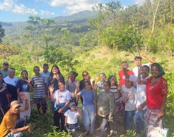 A group of farmers and visitors stand together in a hillside agroforestry field, smiling and waving, with green mountains and trees in the background.
