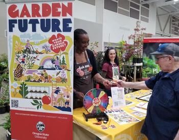 Master Gardener volunteer staffs a “Garden Future” booth, handing materials to a visitor beside a colorful display and information table.