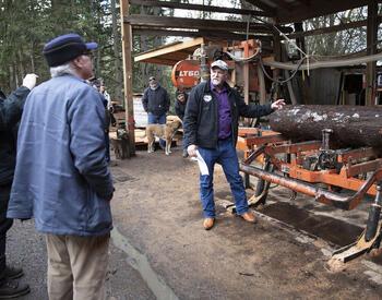 Man stands by log machine outside during Tree School