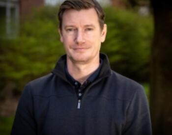 James Osborne is wearing a dark quarter-zip sweater stands outdoors with trees and campus buildings blurred in the background.
