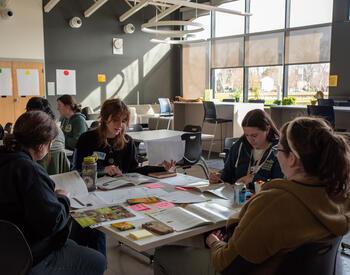 Workshop participants sit around a table in a bright classroom, reviewing worksheets and discussing materials during a hands-on training activity.
