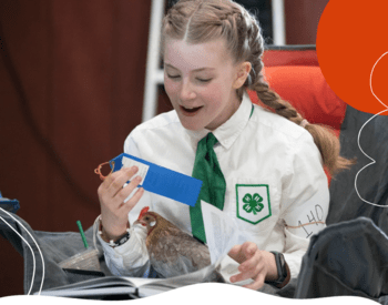 Girl in 4-H uniform with blonde hair in braids. Girl is sitting in lawn chair, holding a blue ribbon in her right hand, with a chicken in her lap. She is also holding a book in her lap. On her left arm a paper with the exhibitor number "140" is rubber banded to her arm, she also has a watch on her left wrist.