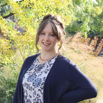 Brunette woman with curly hair wearing a dark blue cardigan over a cream blouse with blue flowers. Wearing a gold chain necklace with her hand on her hip.