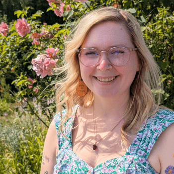 Jen, a blonde woman wearing glasses, smiles in front of a pink rose bush