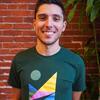 Daniel Wear is a younger white man with a short haircut, wearing a sustainable northwest t-shirt and smiling at the camera, standing in front of a brick wall. The SNW logo consists of abstract mountains, water, and sun in horizontal rainbow colors.