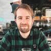 Patrick Shults is sitting at a countertop with an urban background. He is smiling at the camera, wearing a plaid shirt.